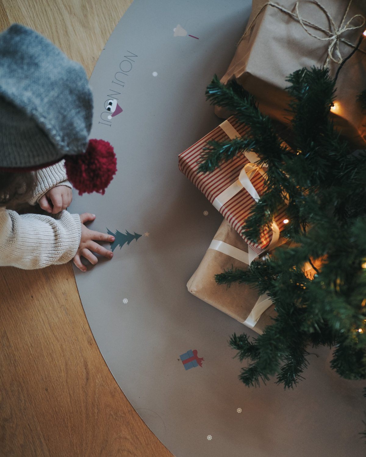 A child sitting next to a fir tree opening presents on our round vinyl floor mat with a Christmas theme, featuring holiday icons such as snowflakes, a Christmas tree, and angel.