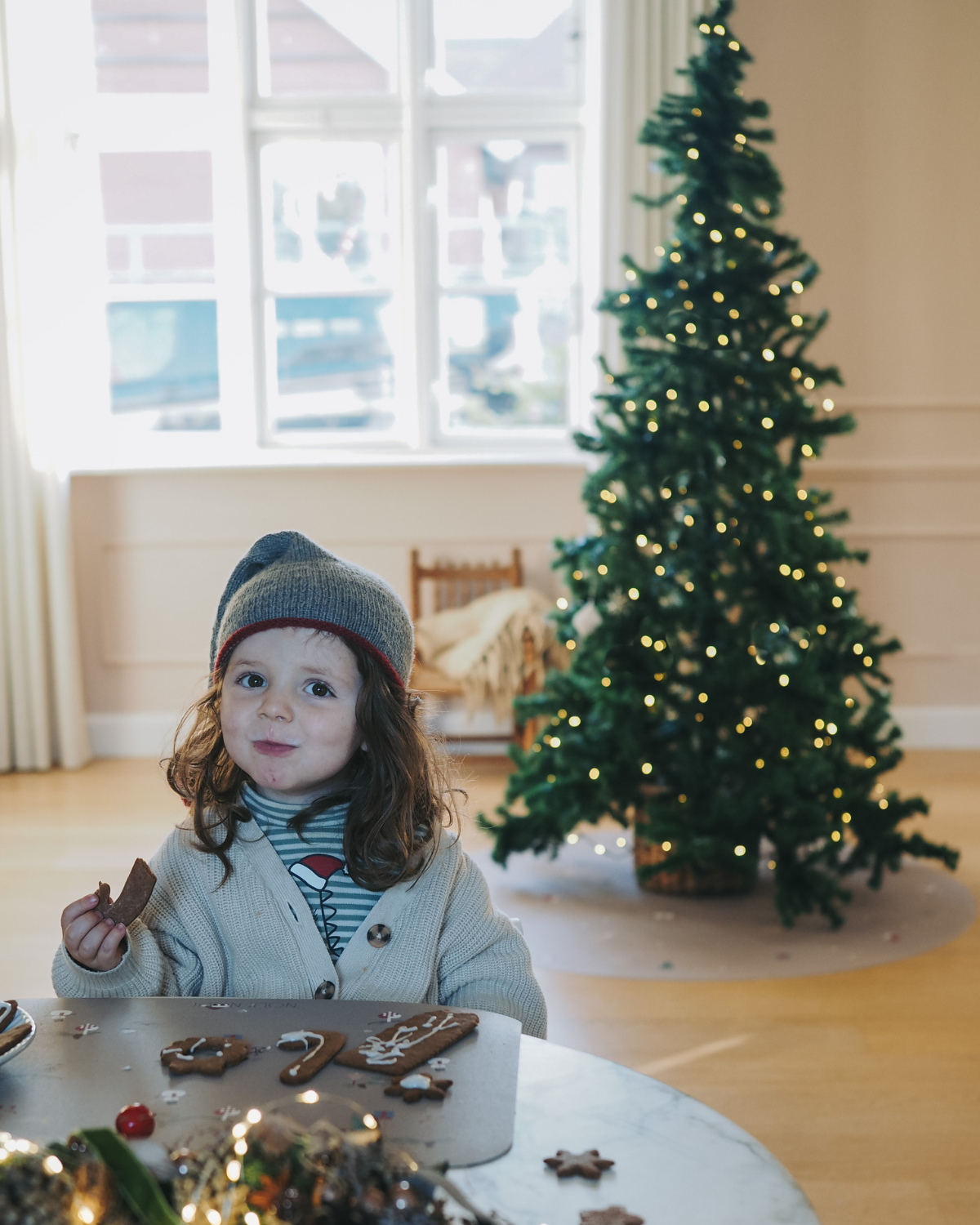A child sitting at the table. Behind is a Christmas tree on our round vinyl floor mat with a Christmas theme, featuring holiday icons such as snowflakes, a Christmas tree, and angel.
