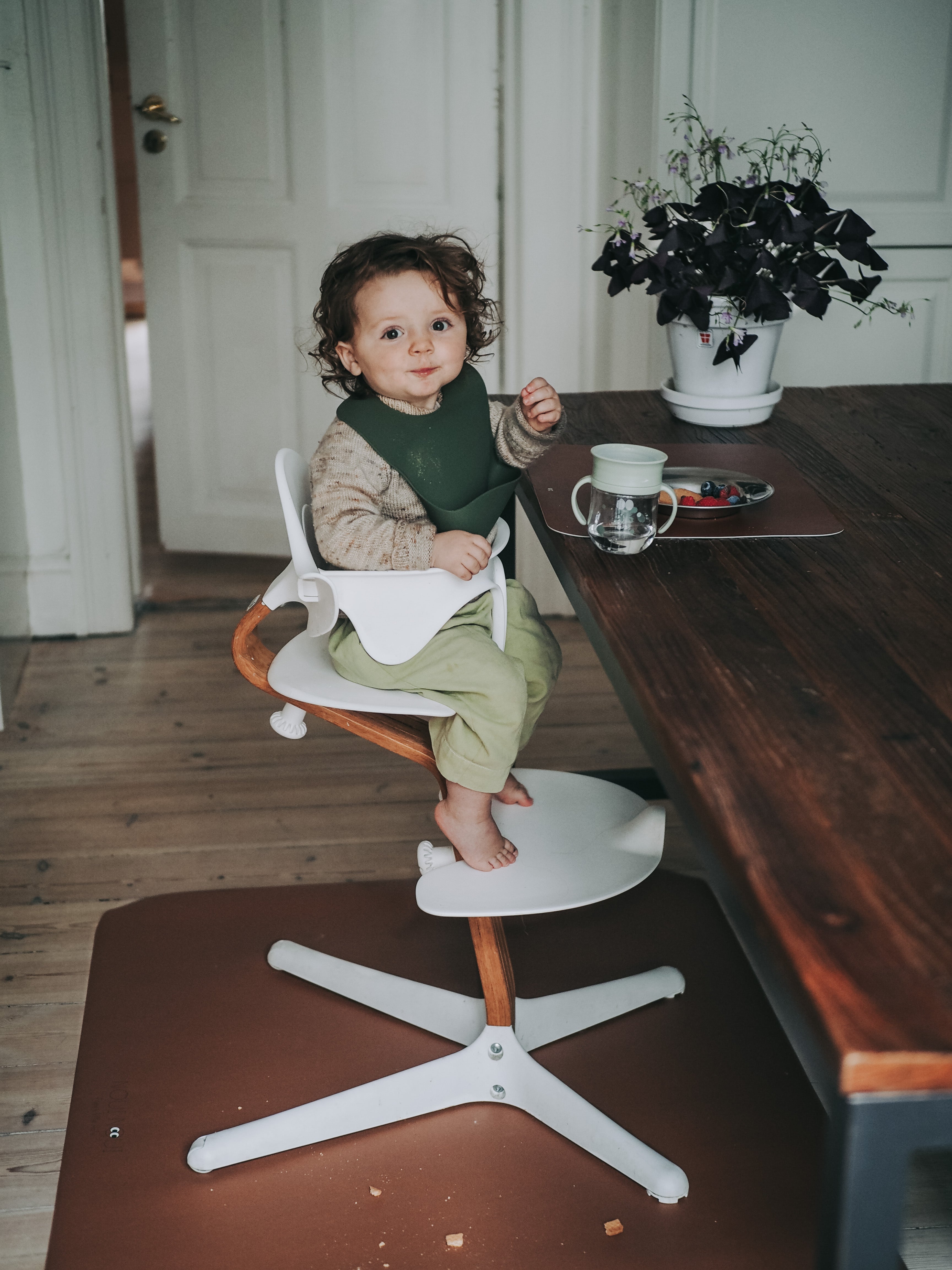 A child wearing our dusty olive silicone bib with adjustable straps and a pocket in the front. Sitting on a high chair.