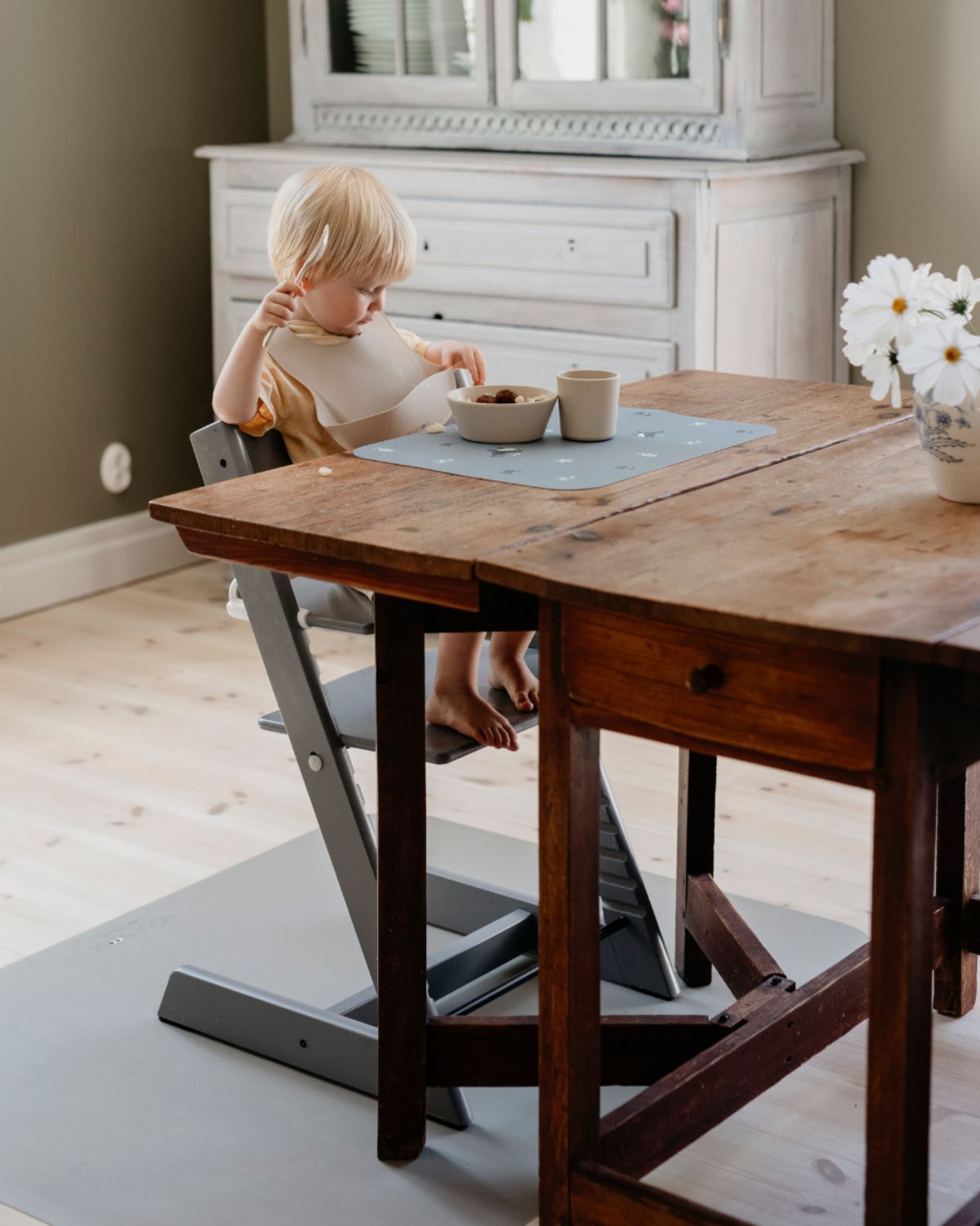 A high chair floor mat made of soft vinyl, in the color olive haze gray. A rectangular mat to protect all kind of floors from spills. Put it under the high chair. Easy to clean.