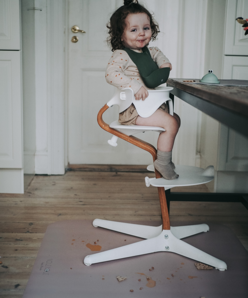 A child wearing our dusty olive silicone bib with adjustable straps and a pocket in the front. Sitting on a high chair.