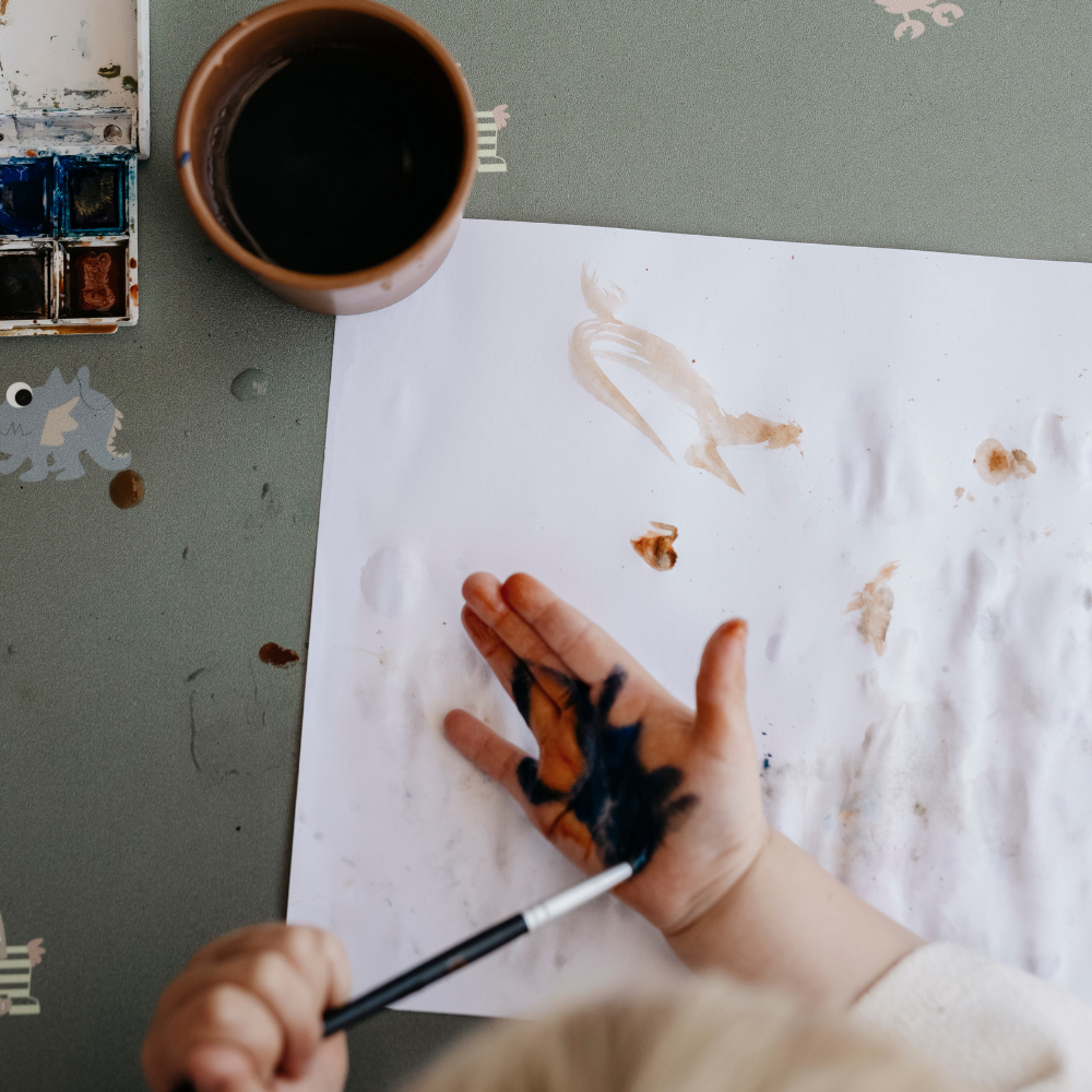 Child painting on our Placemat with dusty olive background featuring a playful pattern of various small, friendly fantasy animal fable icons.