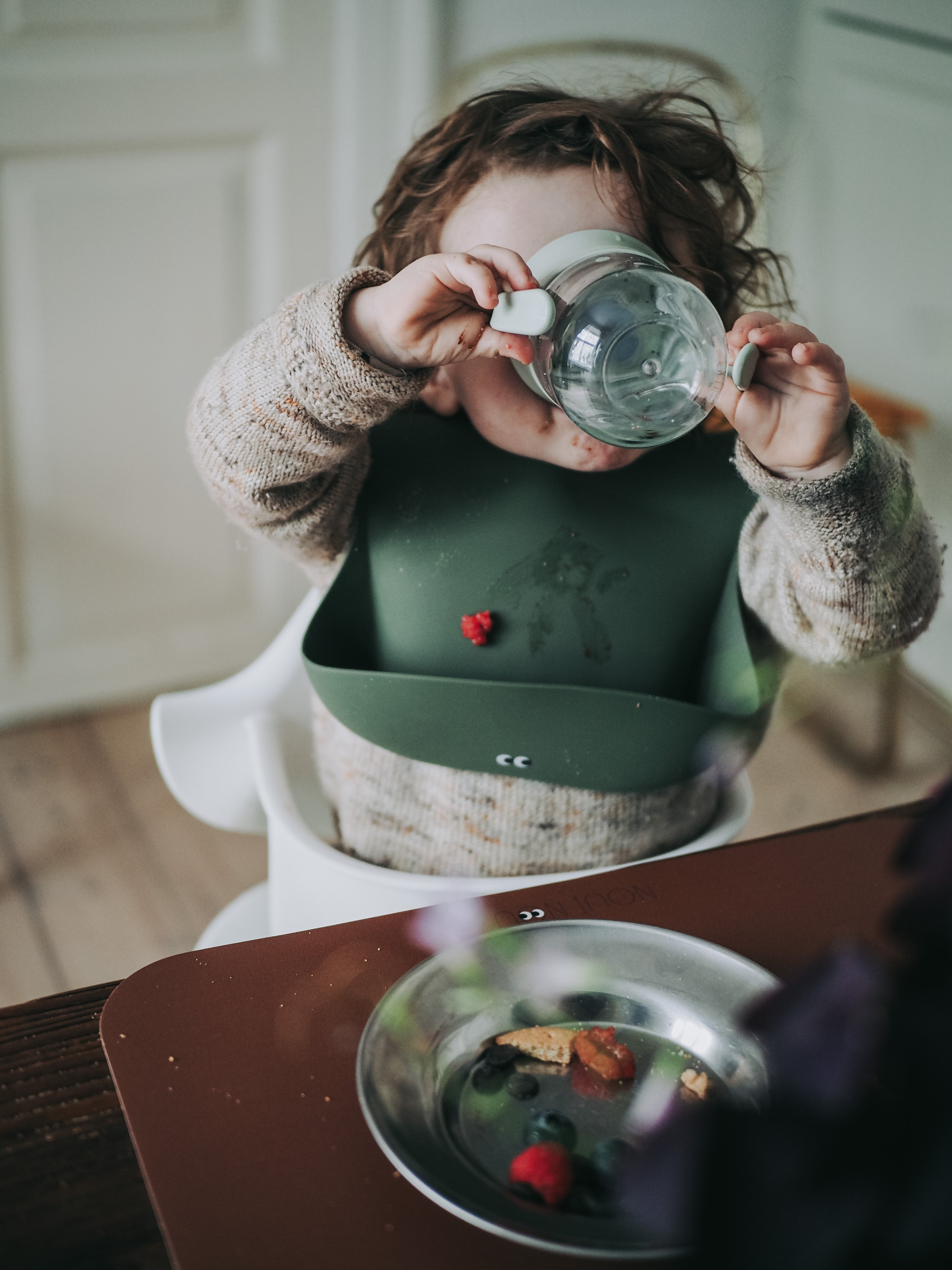 A child drinking while wearing our dusty olive silicone bib with adjustable straps and a pocket in the front