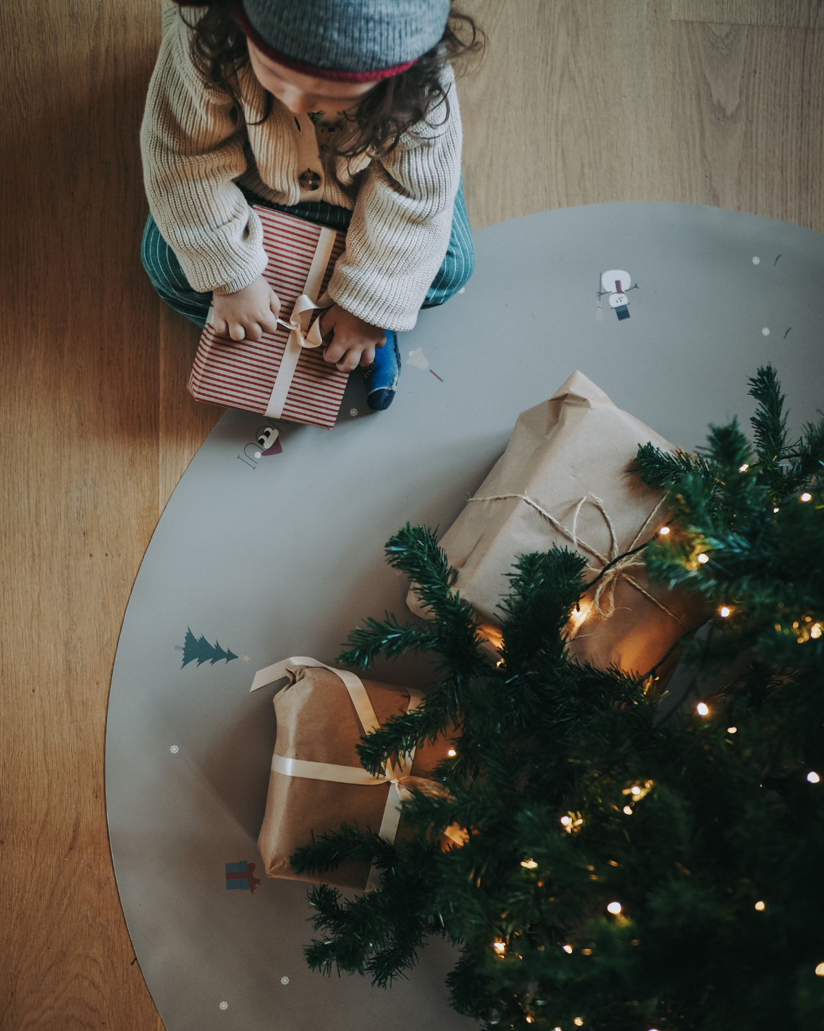 A child sitting next to a fir tree opening presents on our round vinyl floor mat with a Christmas theme, featuring holiday icons such as snowflakes, a Christmas tree, and angel.