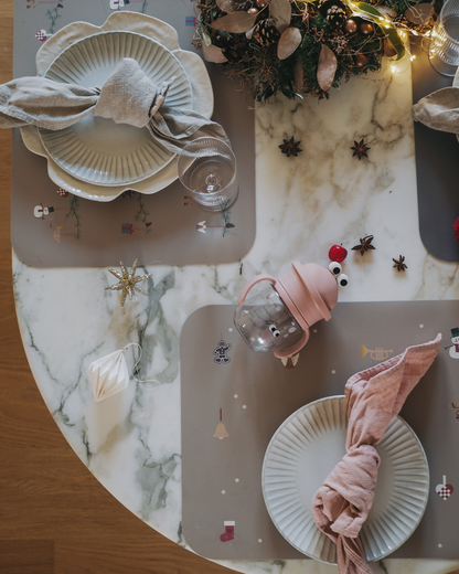 A placemat with a winter-themed design featuring snowflakes, Santa Claus, and Christmas-related icons on a olive haze gray background on a table setting.