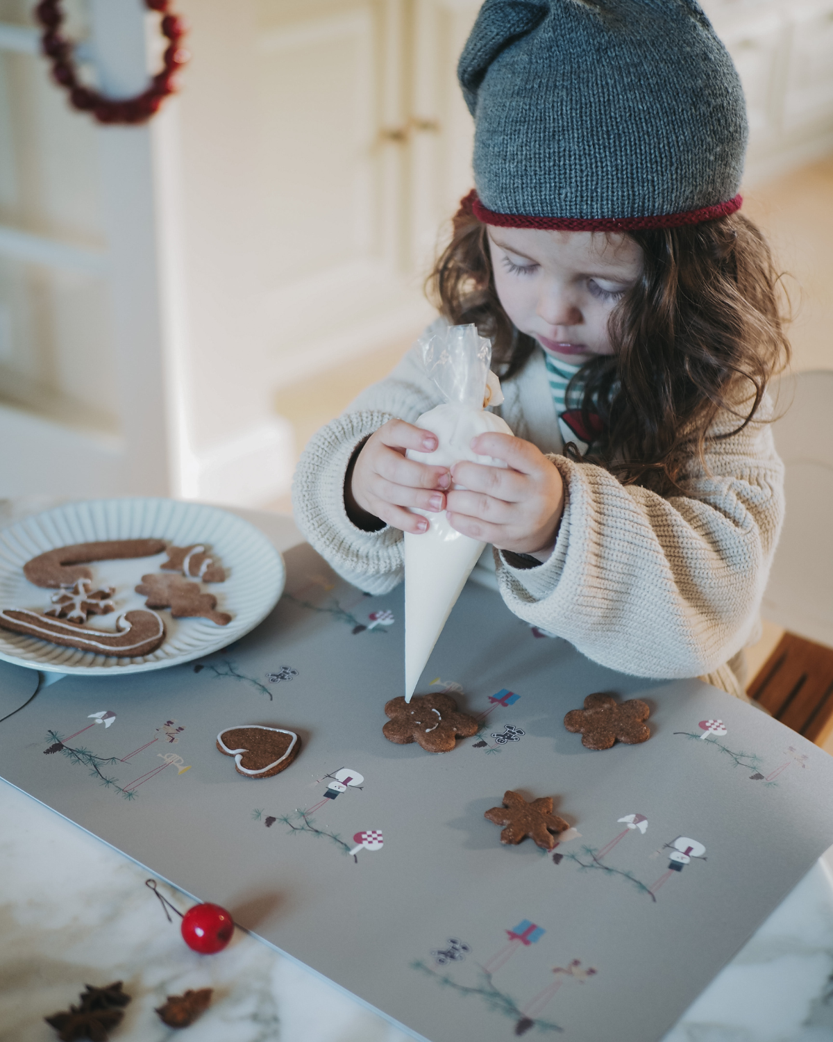 A child decorating cookies on our olive haze gray placemat with Christmas-themed decorations including snowmen, hearts, angel on fir branch.
