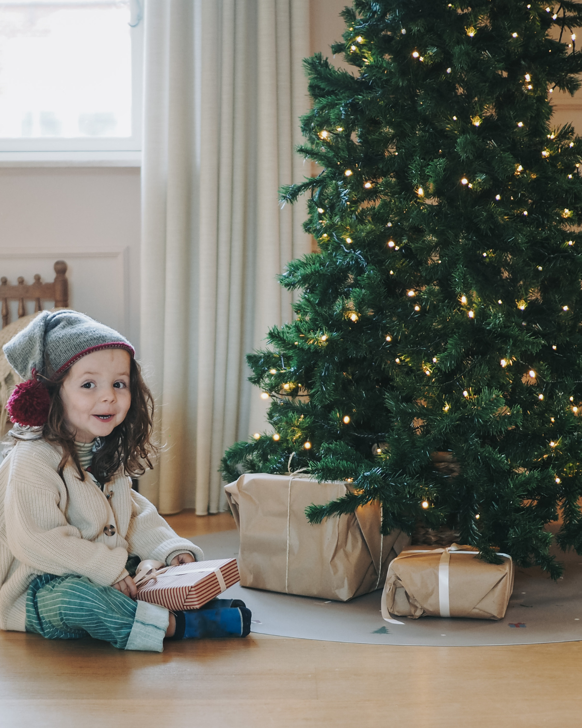A child sitting next to a fir tree opening presents on our round vinyl floor mat with a Christmas theme, featuring holiday icons such as snowflakes, a Christmas tree, and angel.