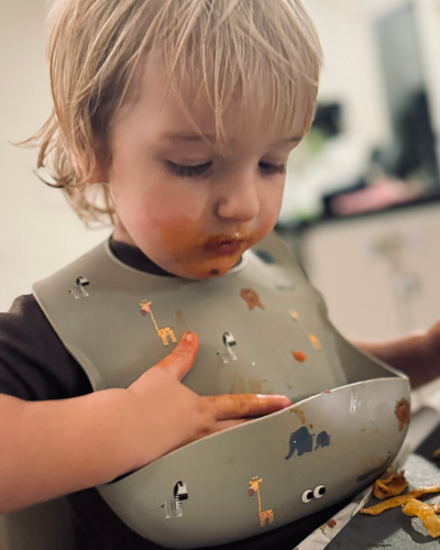 A child wearing our soft silicone bib in olive haze gray color with various small animal illustrations and an adjustable neck strap.