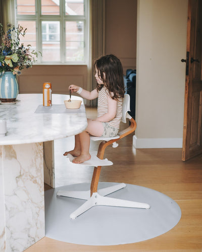 A grey powder learning placemat with illustrations of a plate, fork, knife, and spoon on it. Paired with a round grey powder floor mat under a child high chair. 