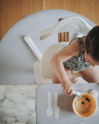 A grey powder learning placemat with illustrations of a plate, fork, knife, and spoon on it. Paired with a round grey powder floor mat under a child high chair. 