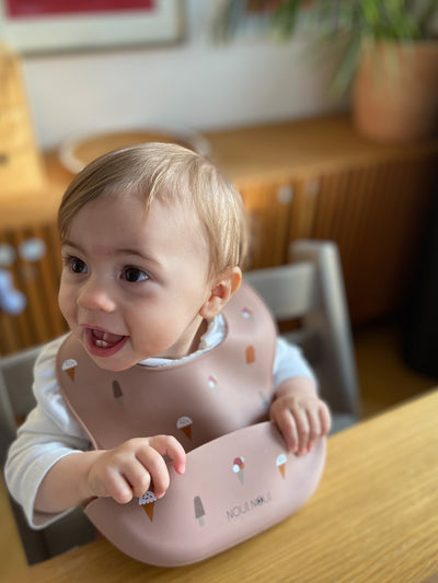 A child wearing our silicone bib in dusty rose color with a pattern of different cute ice cream. The bib has an adjustable strap and an extra large cup to catch food and fluids. 