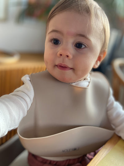 A child sitting at the table wearing our nude silicone bib with adjustable straps and an extra cup to catch food and fluids. 