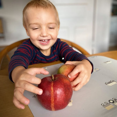 Child playing with an apple on our Placemat in grey powder color with a pattern of cars and other vehicles in grey tones.