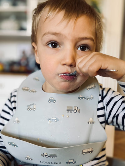 A  child wearing our silicone bib with a pattern of small cute cars and trucks. Adjustable strap in the neck and an extra large cup to catch food an fluids.