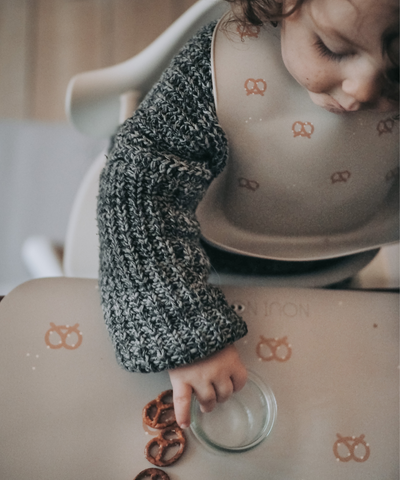 Placemat in nude tone with a pattern of pretzels and salt wearing our nude silicone bib with pretzels.