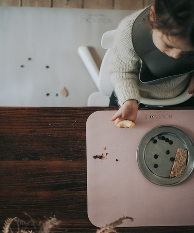A child sitting at the table wearing our warm gray soft silicone bib with adjustable straps and a large cup for catching food.