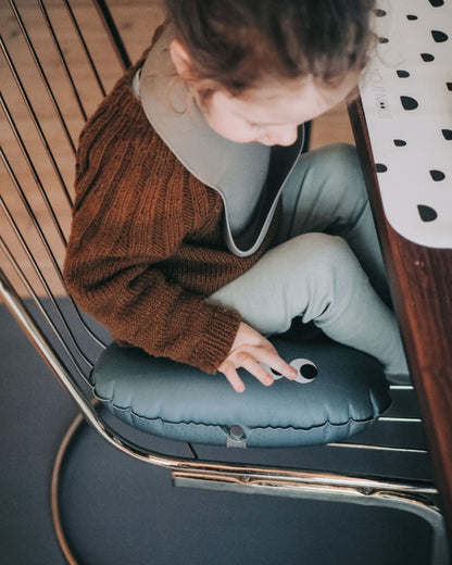 alt="Inflatable ergonomic booster cushion for children with elastic strap and anti-slip backing, placed on a dining chair"