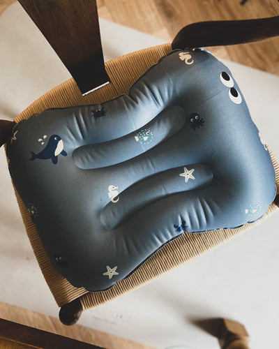 alt="Inflatable ergonomic booster cushion for children with elastic strap and anti-slip backing, placed on a dining chair"