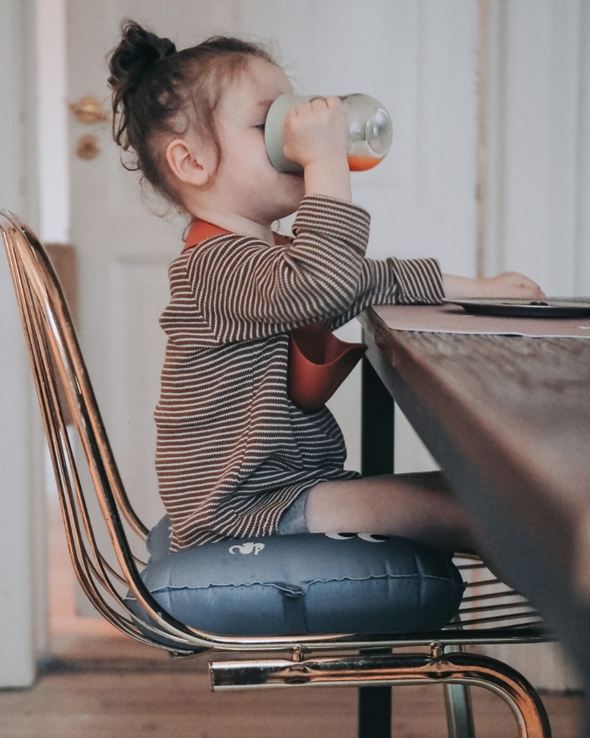 alt="Inflatable ergonomic booster cushion for children with elastic strap and anti-slip backing, placed on a dining chair"