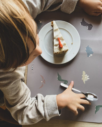 A child eating cake on our Placemat with various illustrations of dinosaurs in shades of green, brown, and gray, on a cocoa background.