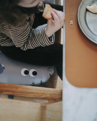 alt="Inflatable ergonomic booster cushion for children with elastic strap and anti-slip backing, placed on a dining chair"