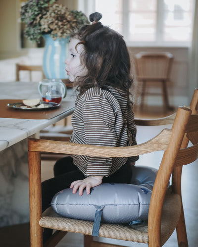 alt="Inflatable ergonomic booster cushion for children with elastic strap and anti-slip backing, placed on a dining chair"