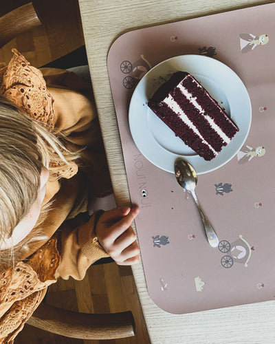 A child eating cake on our placemat with a princess-themed pattern including illustrations of princesses, castles, and hearts in pink tones.