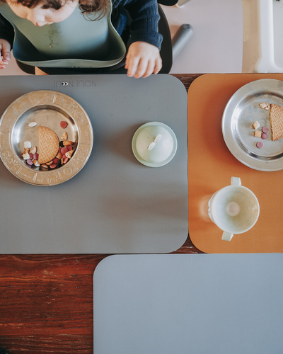 A dusty blue placemat made of soft vinyl displayed together with other placemats. 
