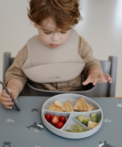 A child sitting at the table wearing our nude silicone bib with adjustable straps and an extra cup to catch food and fluids. 