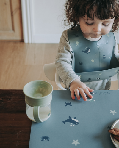 A child wearing our soft silicone bib in dusty blue color with small see animal illustrations and an adjustable neck strap. Paired with our placemat in dusty blue.