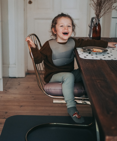 A child sitting at the table wearing our warm gray soft silicone bib with adjustable straps and a large cup for catching food.