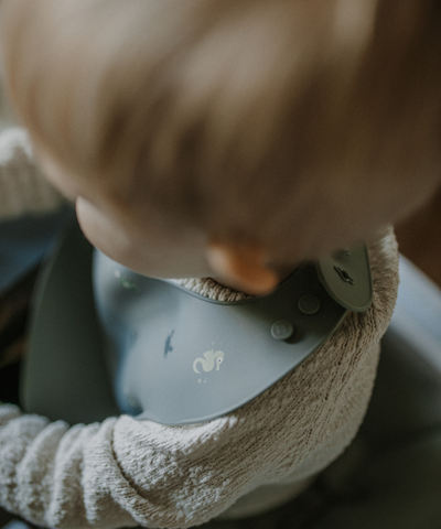 A child wearing our soft silicone bib in dusty blue color with small see animal illustrations and an adjustable neck strap.